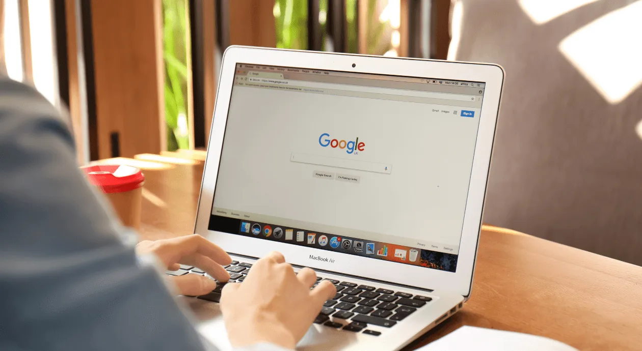 A white person's hand typing on an open laptop showing the Google homepage, on a wooden table in a sunlit room.