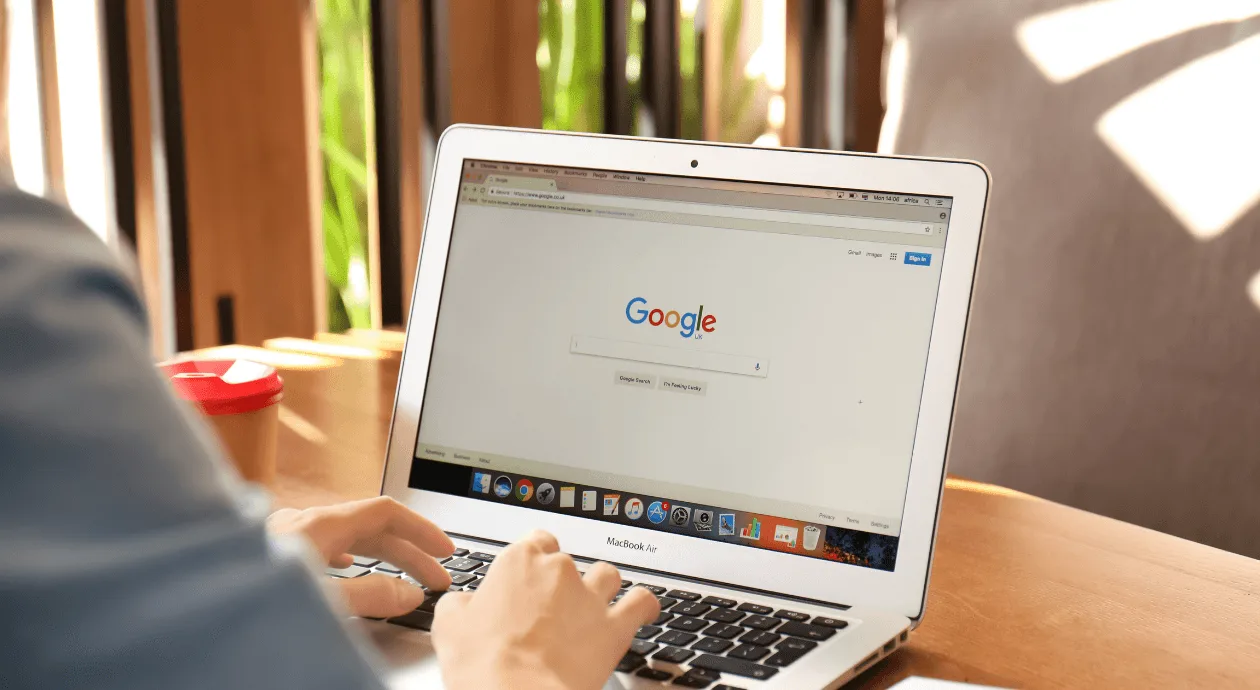 Person typing on a laptop with the Google homepage open on the screen, on a wooden desk with a cup of coffee beside it.