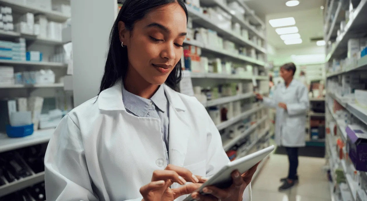 Pharmacist using a tablet to manage the pharmacy's Google Business Profile in an aisle lined with medicine shelves.